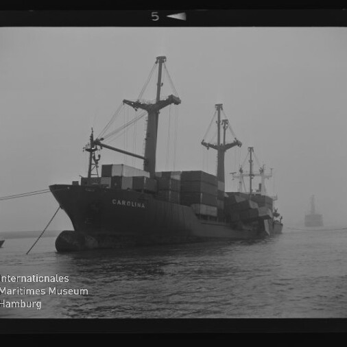 Containerschiff Carolina mit gestapelten Containern und Ladekränen fährt in nebligem Hafen, Museumsemblem unten links.