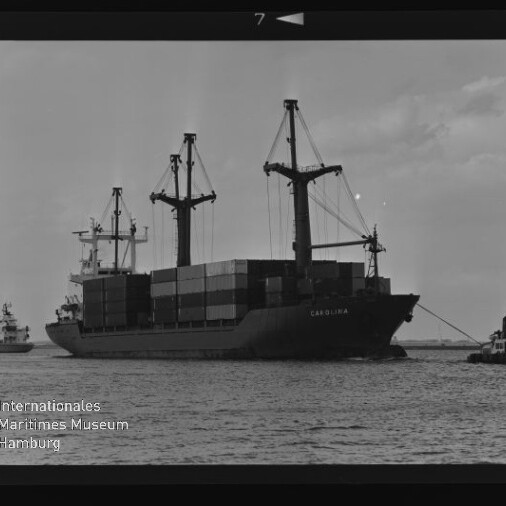 Schwarzweißaufnahme eines Containerschiffs CAROLINA mit drei Ladekränen und gestapelten Containern am Hafen.