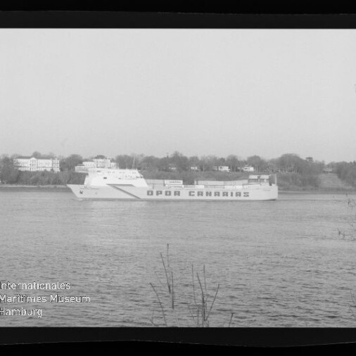 Große Fähre mit auffälligem Schriftzug fährt auf einem Fluss, im Hintergrund Ufer mit Häusern.