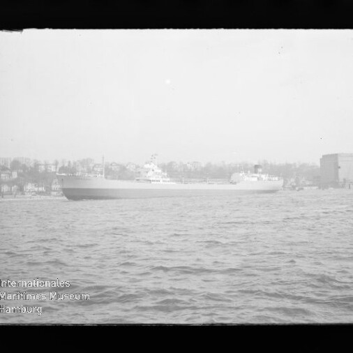 Großes Frachtschiff auf dem Wasser vor Ufer mit Häusern und großem Silo; Museumslogo links unten