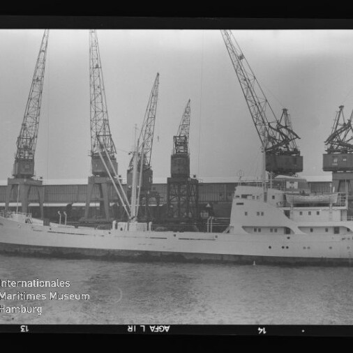 Weißes Frachtschiff am Kai vor mehreren hohen Hafenkränen, schwarzweißes Foto mit Museumsstempel unten links.