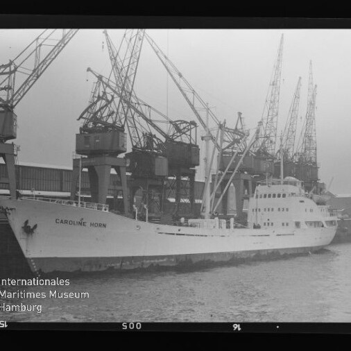 Frachtschiff CAROLINE HORN liegt am Industriequai vor einer Reihe großer Hafenkräne, schwarzweiß.