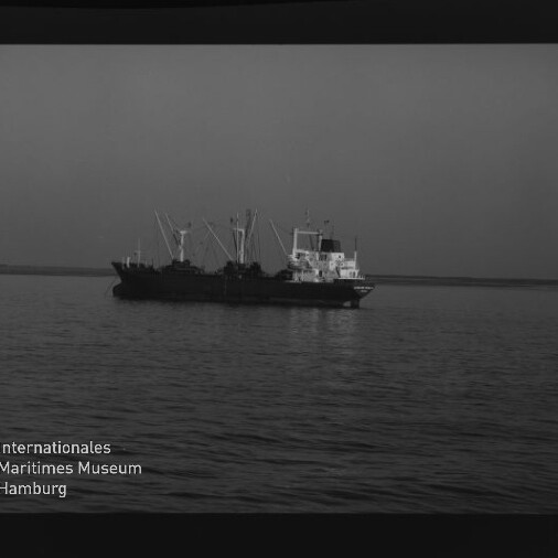 Schwarzweiß-Negativ eines Frachtschiffs mit Ladekränen vor ruhigem Meer und flachem Horizont, Filmrand und Museumslogo sichtbar.