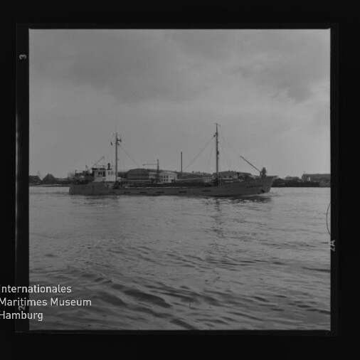 Schwarzweiß-Foto eines Frachtschiffs auf ruhigem Wasser vor Hafenbauten; Wasserzeichen 'Internationales Maritimes Museum Hamburg'.