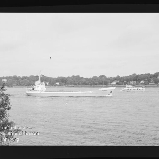 Frachtschiff fährt auf ruhigem Fluss vor baumbewachsener Uferlinie mit kleinem Ausflugsschiff und Museumswasserzeichen.