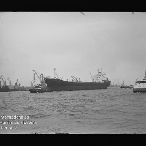 Großes Frachtschiff im Hafen, begleitet von Schleppern, mit Krananlagen und weiteren Schiffen im Hintergrund.