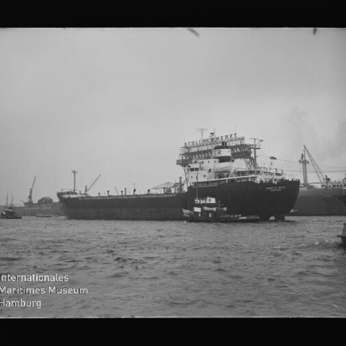 Großes Frachtschiff mit Schlepper neben Bug im Hafen, Kräne im Hintergrund und Museumslogo unten links.