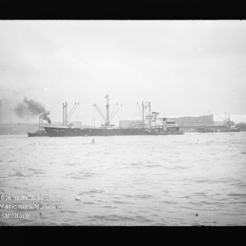 Frachtschiff mit rauchendem Schornstein und Ladekränen vor Hafenanlagen, Wasser im Vordergrund, Wasserzeichen unten links.