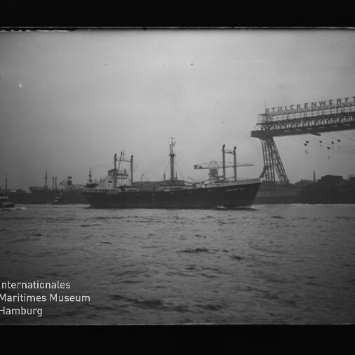 Frachtschiff vor Industriehafen mit großem Portalkran und sichtbarem Schriftzug und Museumslogo unten links.