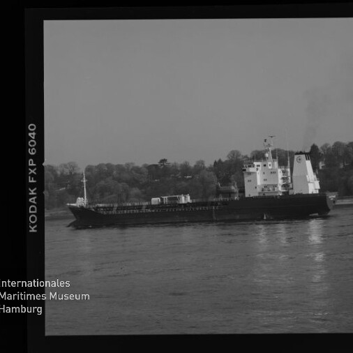 Schwarzweißfoto eines Frachtschiffs auf ruhigem Wasser vor bewaldetem Ufer, mit Filmrand (KODAK FXP 6040) und Museumslogo.