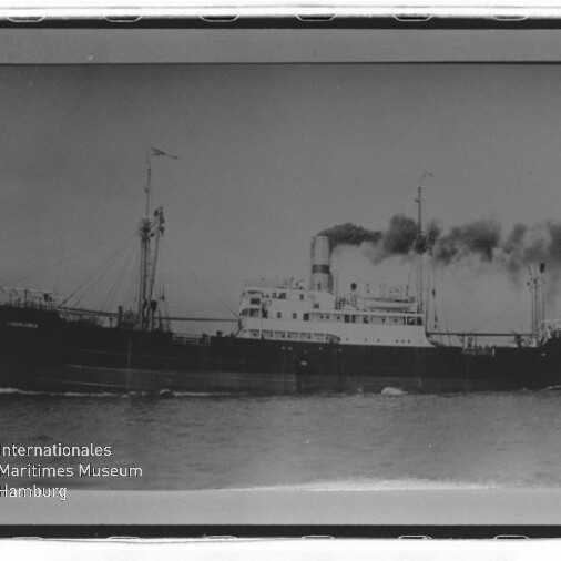 Schwarzweißfoto eines Dampfschiffs auf See mit Rauch aus dem Schornstein und Ladegeschirr; Museumslogo links unten.