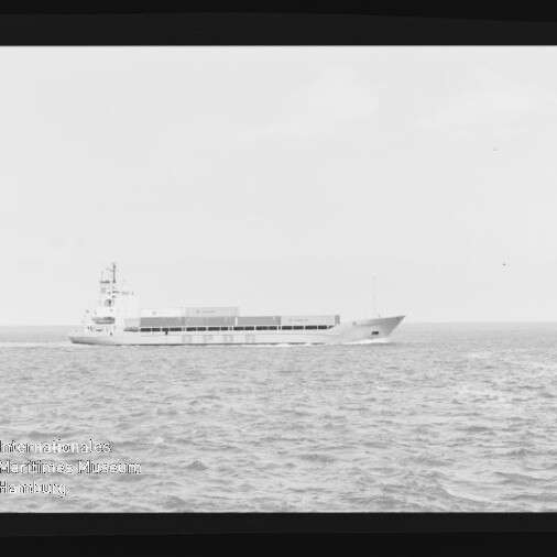 Schwarzweißaufnahme eines niedrigen Frachtschiffs mit Containern auf ruhiger See, seitliche Ansicht mit Filmrand und Museumslogo.