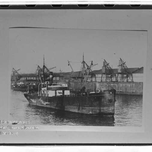 Frachtschiff im Hafen vor Lagerhäusern und Kränen in Schwarzweiß, mit Wasserzeichen Internationales Maritimes Museum Hamburg.