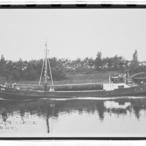 Kleines Frachtschiff mit Mast auf ruhigem Wasser, Spiegelung und Baumreihe im Hintergrund; Museumswasserzeichen.