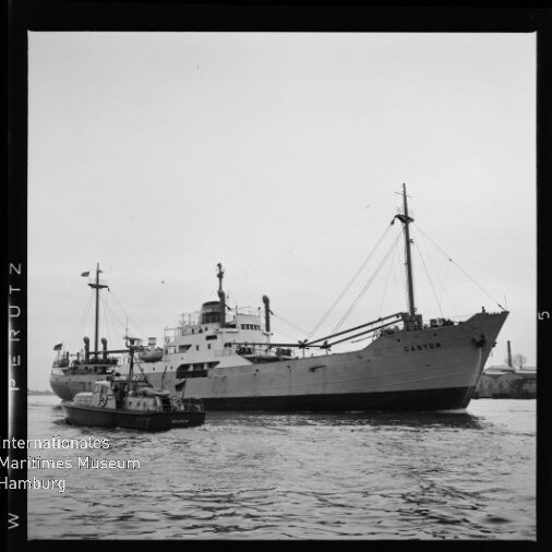 Schwarzweiß-Foto eines Frachtschiffs mit Masten und Ladebäumen, begleitet von einem Schlepper im Hafen.