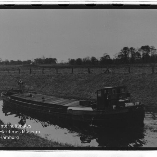 Schwarzweißaufnahme eines langen Lastkahns auf einem Kanal mit kleinem Steuerhaus und flachem Deck, Ufer mit Bäumen im Hintergrund.