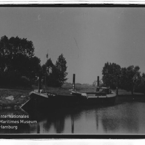 Schwarzweißfoto eines am Ufer vertäuten Lastkahns vor Baumreihe, ruhige Wasserfläche mit Spiegelung und Museumslogo links unten.