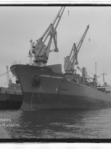 Großes Frachtschiff mit mehreren Ladekränen am Kai in einer Schwarzweiß-Hafenaufnahme.
