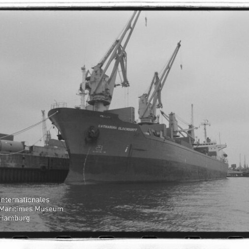 Großes Frachtschiff mit mehreren Ladekränen am Kai in einer Schwarzweiß-Hafenaufnahme.