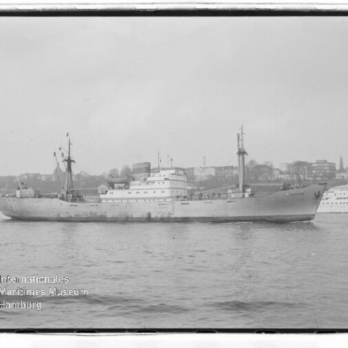 Schwarzweißaufnahme eines Frachtschiffs auf ruhigem Wasser vor einer Stadtkulisse; links unten Wasserzeichen Internationales Maritimes Museum Hamburg