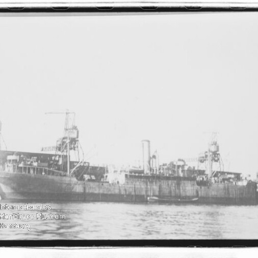 Langes, altes Frachtschiff mit Deckkränen und mittigem Schornstein am Kai, ruhiges Wasser im Vordergrund.