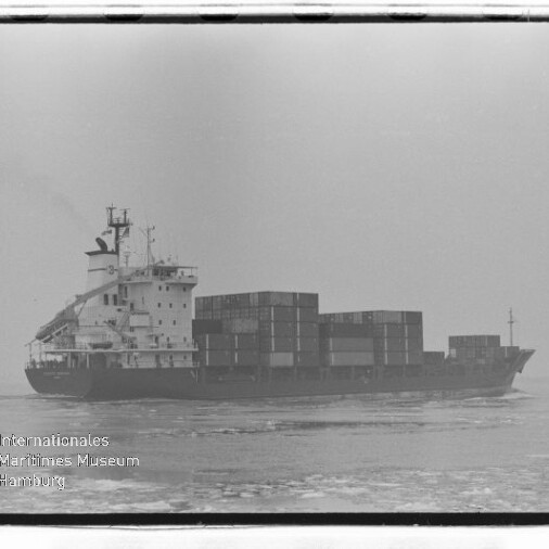 Containerschiff mit gestapelten Containern fährt bei nebligem Himmel über leicht eisbedecktes Meer; Museumslogo unten links.