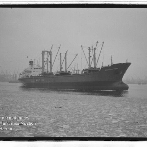Frachtschiff mit mehreren Ladekränen liegt in einem nebligen Hafen, Eisschollen bedecken das Wasser.