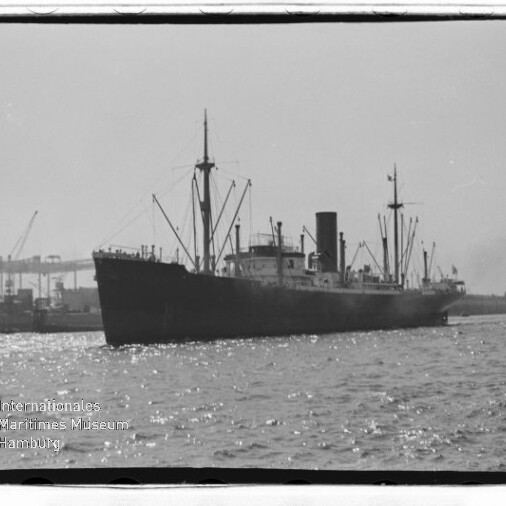 Schwarzweiß-Foto eines Frachtschiffs mit Schornstein und Ladebäumen im Hafen, im Hintergrund Hafenkräne und Museumswasserzeichen.