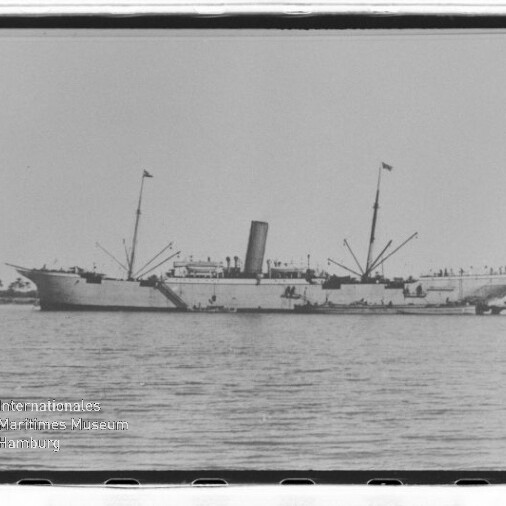 Dampfschiff mit Schornstein und zwei Masten, mit Fahnen auf ruhigem Wasser vor Küste; kleiner Schlepper daneben.