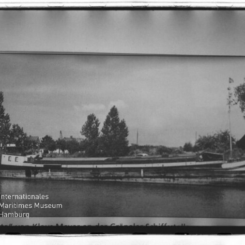 Schwarzweiß-Foto eines langen Binnenschiffs am Ufer vor Bäumen und Häusern, gerahmt; Schriftzug Internationales Maritimes Museum