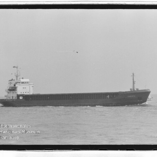 Schwarzweißaufnahme eines Frachtschiffs auf ruhiger See, mit kleinem Segelboot rechts und Museumswasserzeichen links unten.