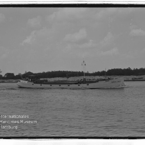 Schwarzweißfoto eines langen Küstenmotorschiffs auf ruhiger See vor baumbestandener Küste; Wasserzeichen 