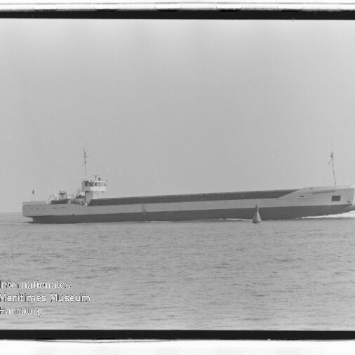 Langes, flaches Frachtschiff mit kleinem Aufbau fährt ruhig auf offener See, neben dem Bug eine Boje.