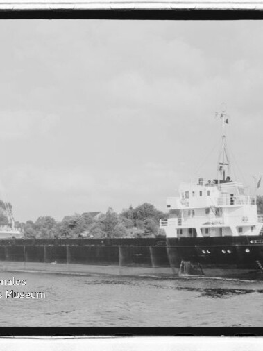 Schwarzweißfoto eines Frachtschiffs auf einem Kanal mit bewaldetem Ufer; Schriftzug Internationales Maritimes Museum Hamburg links unten.