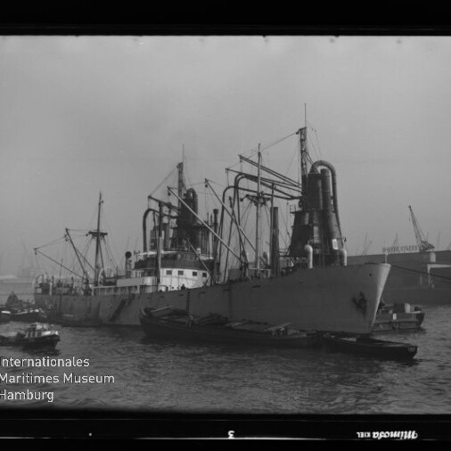 Schwarzweißfoto eines großen Frachtschiffs mit hohen Schornsteinen und Kränen im Hafen, kleine Begleitboote am Bug.