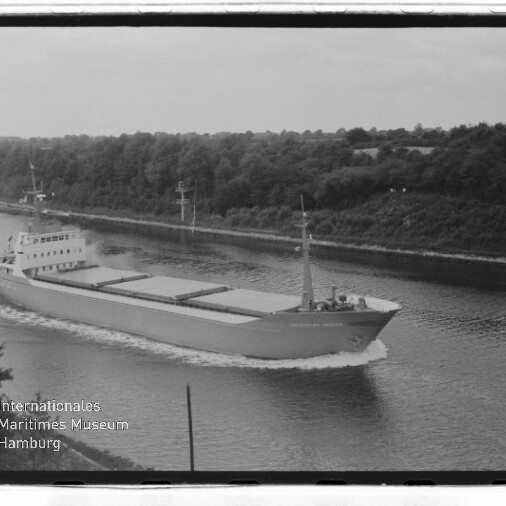 Frachtschiff fährt durch Kanal mit bewaldetem Ufer, links Schriftzug Internationales Maritimes Museum Hamburg.