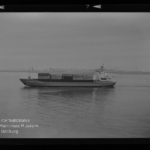 Containerschiff in Seitenansicht auf ruhiger See mit gestapelten Containern und schwacher Hafen-Silhouette am Horizont.