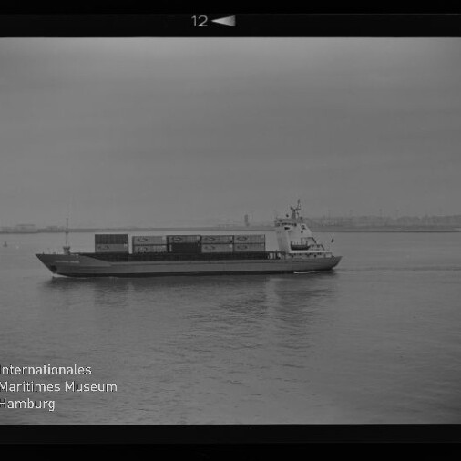 Containerschiff seitlich auf ruhiger See mit auf Deck gestapelten Containern und flachem Küstenhorizont