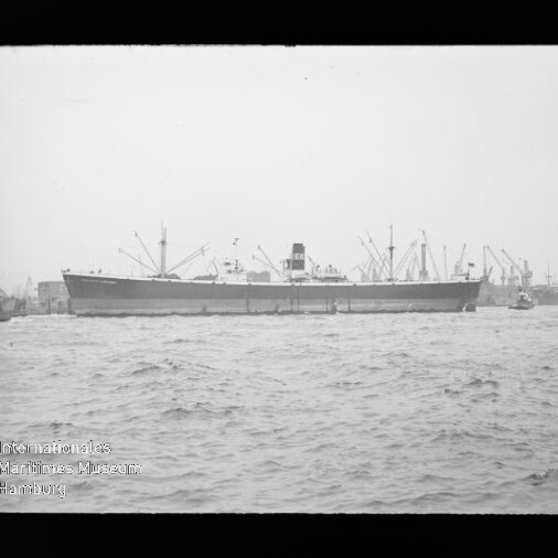 Großes Frachtschiff an der Kaimauer mit Hafenkränen im Hintergrund und unruhiger See im Vordergrund.