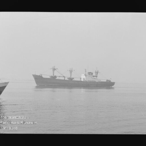 Schwarzweißaufnahme eines Frachtschiffs mit Ladekränen auf ruhiger, nebliger See; links der Bug eines zweiten Schiffs und das Wasserzeichen des Internationalen Maritimen Museums Hamburg