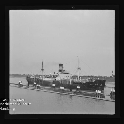 Dampffrachter mit Schornstein und Ladegeschirr am Pier in einer Schwarzweißaufnahme, Schriftzug Internationales Maritimes Museum Hamburg sichtbar
