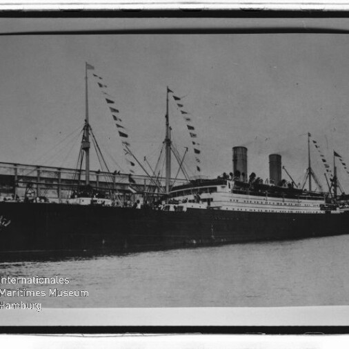 Zweischornsteiniges Passagierschiff mit Wimpelflaggen an Masten am Kai, historische Schwarzweißaufnahme.