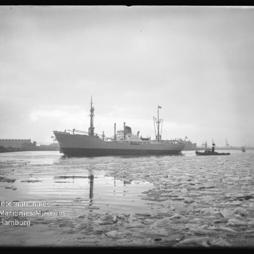 Frachtschiff im Hafen, treibende Eisschollen im Vordergrund und ein Schlepper neben dem Schiff.