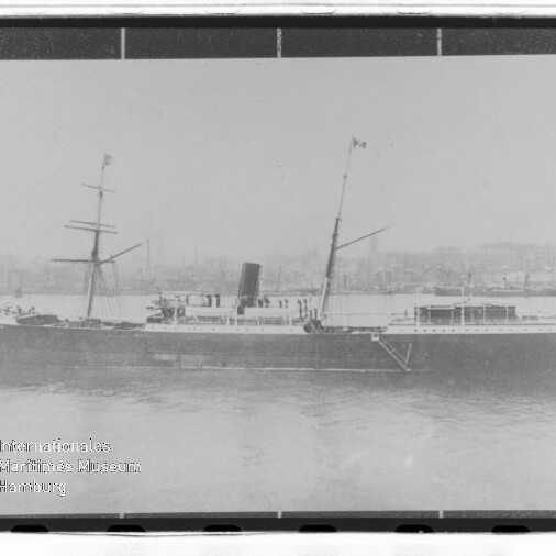 Schwarzweiß-Seitenansicht eines Dampfschiffs mit Schornstein und Masten im Hafen, Stadt im Hintergrund und Museumswasserzeichen links unten