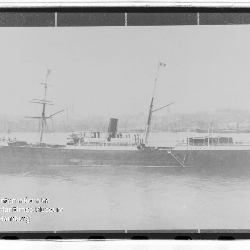 Schwarzweiß-Foto eines historischen Dampfschiffs mit einem Schornstein und zwei Masten, liegend am Hafen vor einer Stadtkulisse.