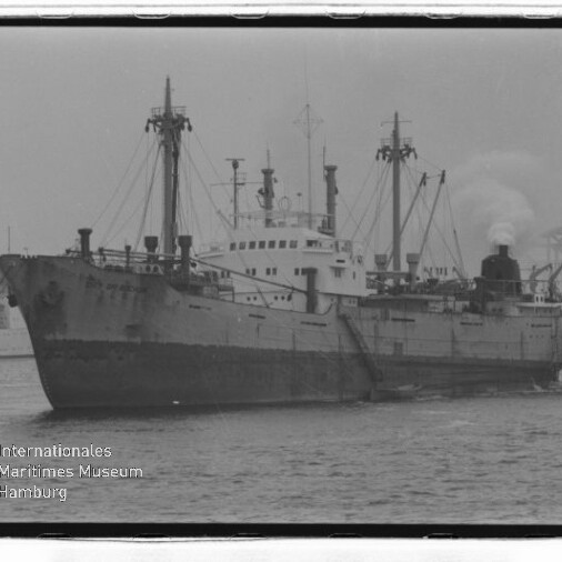 Schwarzweißfoto eines Frachtschiffs mit rauchendem Schornstein und Ladekränen im Hafen, Brücke und Museumswasserzeichen sichtbar.