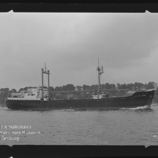 Schwarz-weiß-Foto eines Frachtschiffs mit zwei Ladebäumen und zwei Masten, das vor bewaldeter Küste fährt; Filmrand und Museumslogo sichtbar.