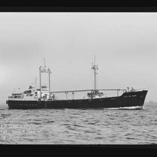 Schwarzweiß-Foto eines Frachtschiffs mit zwei Masten und Ladebäumen, auf ruhiger See fahrend; Museumslogo unten links.