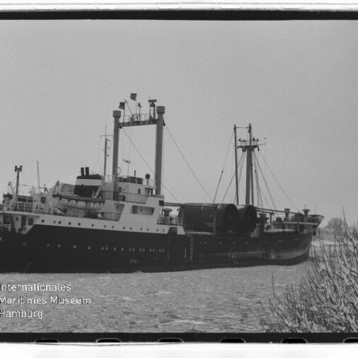 Schwarzweißfoto eines Frachtschiffs mit Masten und großen Rollen an Deck auf ruhigem Wasser; Museumslogo unten links.
