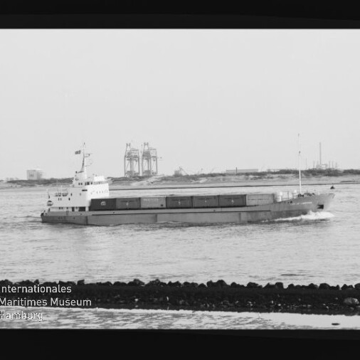 Schwarzweißfoto eines Frachtschiffs vor Hafenkränen; im Vordergrund ein Wellenbrecher; Wasserzeichen des Maritimen Museums Hamburg.
